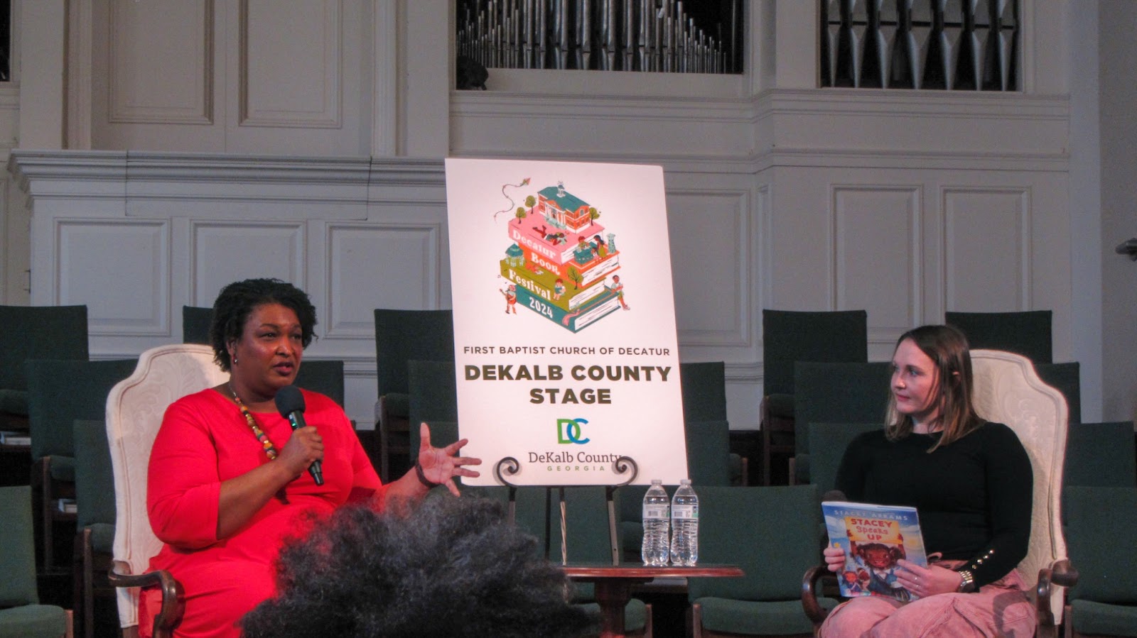 Stacey Abrams sitting down for an interview. Behind Abrams is a poster that reads 'Dekalb County Stage'. Abrams is actively talking in the image.