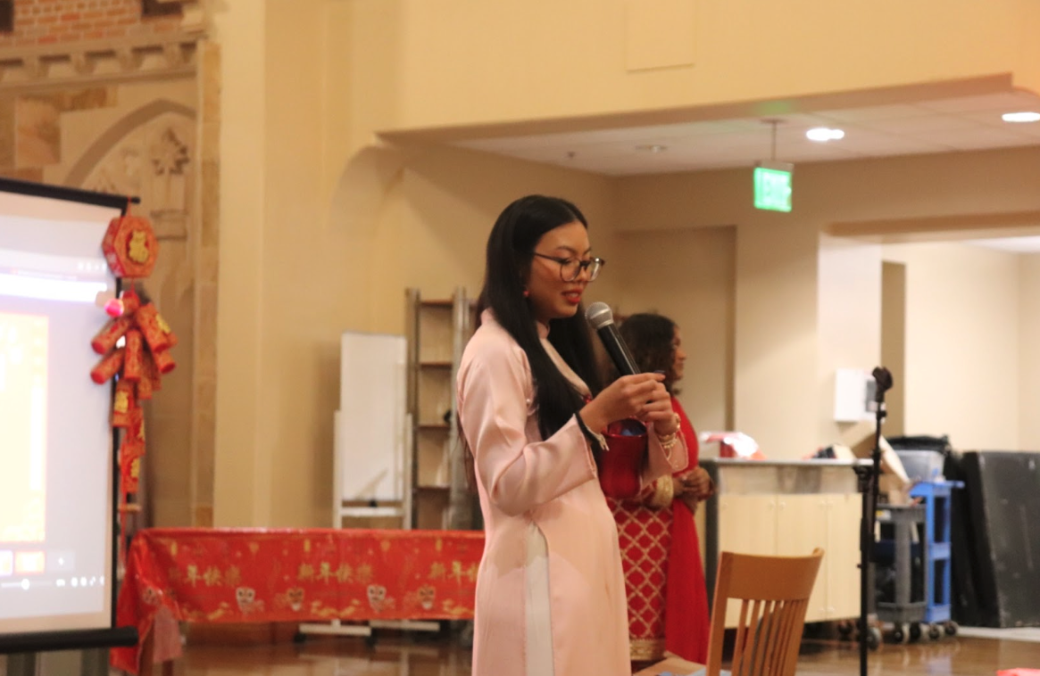 Event MC, Aom Juneau, speaking into a microphone. Their hair is long and worn down, they have black glasses, and they are wearing a pink ao dai.